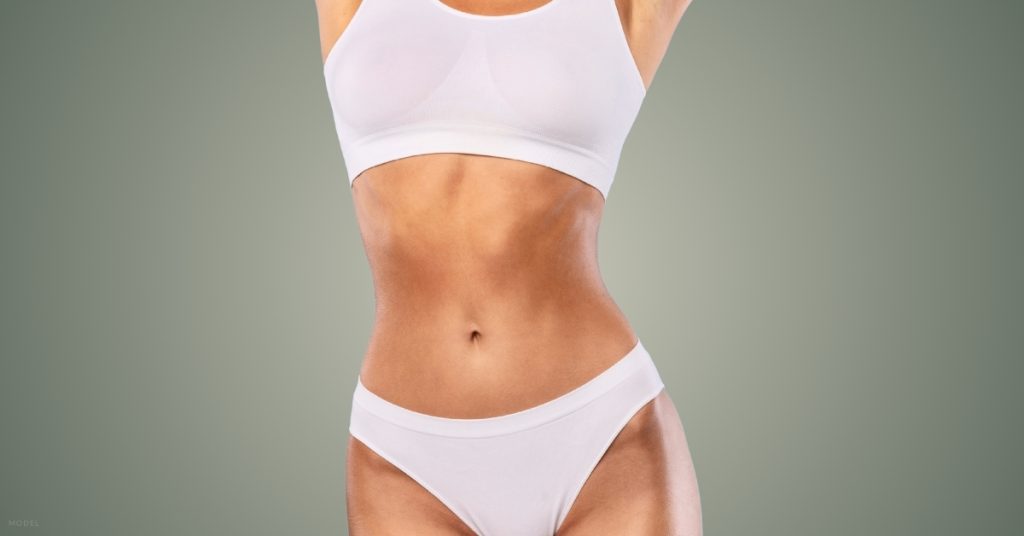 Woman in white undergarments with sculpted stomach and her hands on her stomach and waist. (Model)