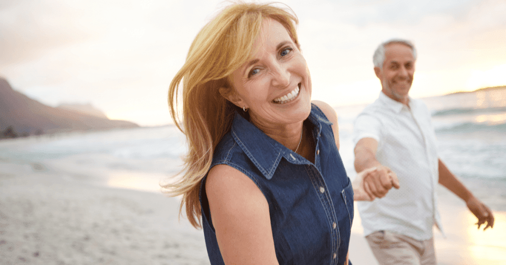 Attractive middle aged woman smiling at beach and holding hands with husband in background
