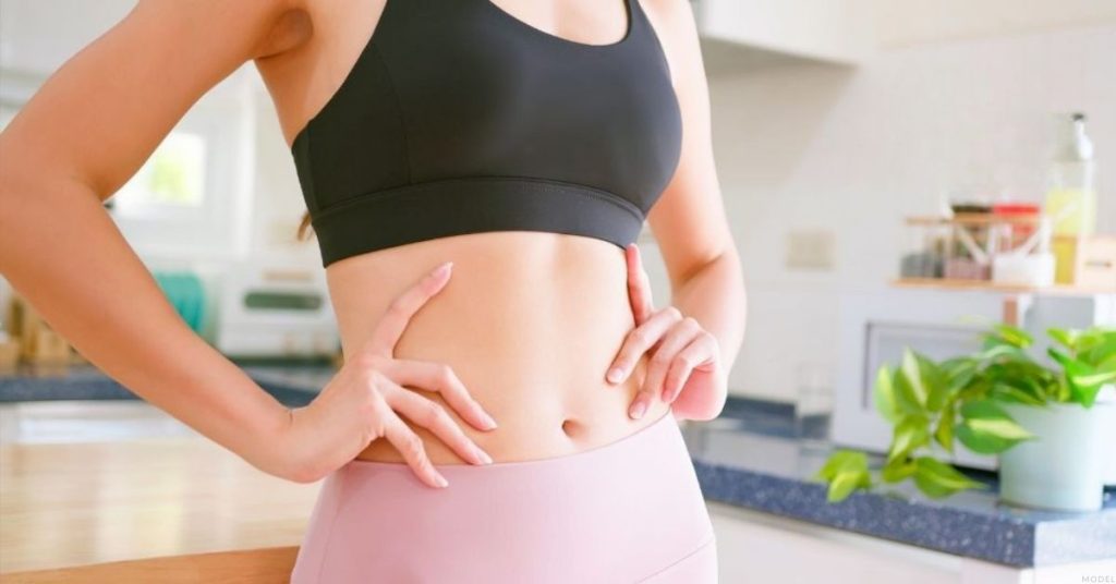 A person is standing in a kitchen with their hands on their stomach (model)