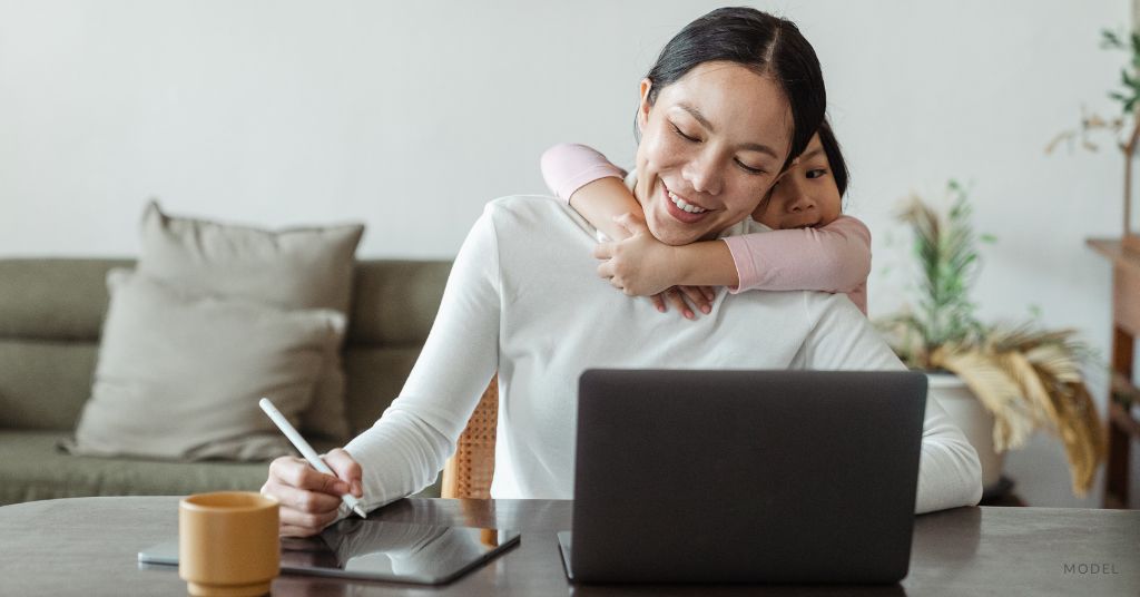 A mother smiling while taking notes as her little girl hugs her from behind. (Model)