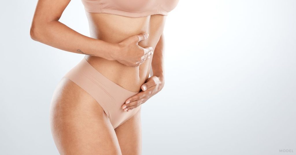 A woman in a nude bra and underwear with their hands on their stomach. (Model)